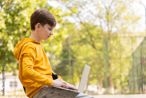 Thoughtful teenager boy resting. Holding and using a laptop for networking on a sunny day, outdoors. photo