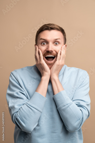 Happy surprised man looking at camera smiling with opened mouth wide spreaded eyes. Portrait amazed crazy guy holding face on beige background. Banner poster placard contains sincere human emotions.  © DimaBerlin