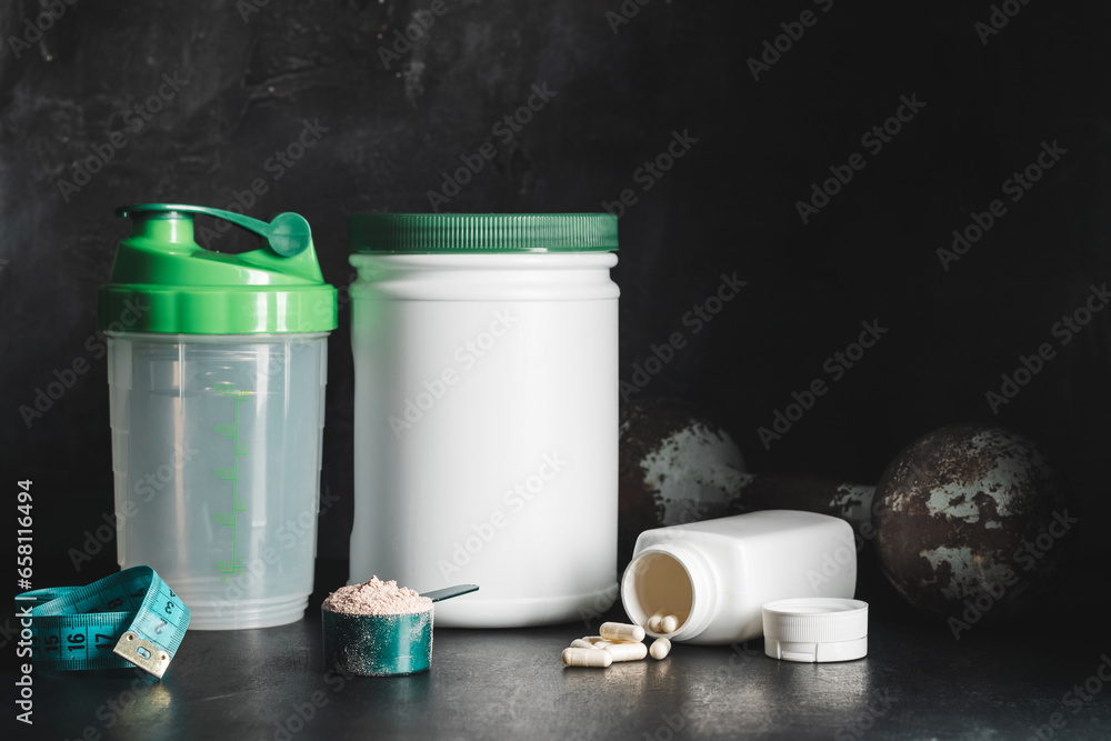Fototapeta premium Chocolate whey protein powder in measuring spoon, white capsules of amino acids and vitamins, measuring tape, old rusty dumbbell, plastic shaker on dark background. bodybuilding food supplements