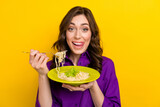 Portrait of excited cheerful person tongue lick teeth hands hold fork spaghetti plate isolated on yellow color background
