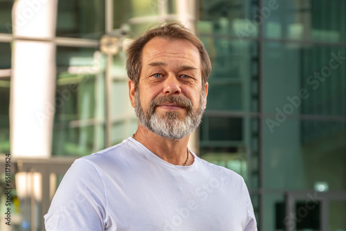 An elderly man 50-55 years old with a beard and an athletic build against the backdrop of modern city buildings, looks at the camera.