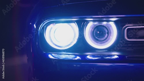 Close up front headlights of muscle car turning on . American sport car is ready to start the race . Engine starting to work . Blue vintage automobile on dark background . Headlamp blinking at night