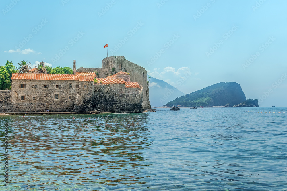 View of the fortress walls with red tiled roofs of the Old Town of Budva and the Island of St. Nicholas in the Adriatic Sea, Montenegro. The Montenegrin flag flutters over the Medieval fortress