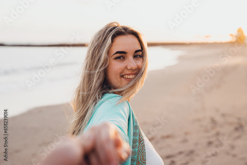 Portrait close up of one young beautiful attractive blonde woman walking on the beach holding boyfriend’s hand looking at the camera. Enjoying free time at vacations concept lifestyle.. #658239014