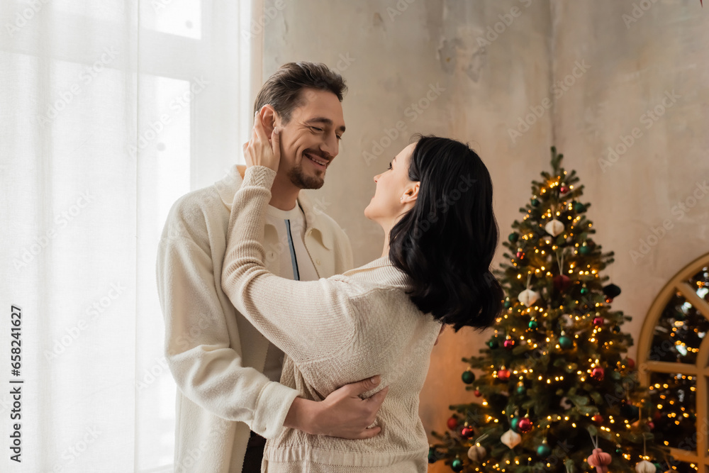 joyful couple in comfortable home wear smiling and hugging near decorated Christmas tree in bedroom joyful couple in comfortable home wear smiling and hugging near decorated Christmas tree in bedroom