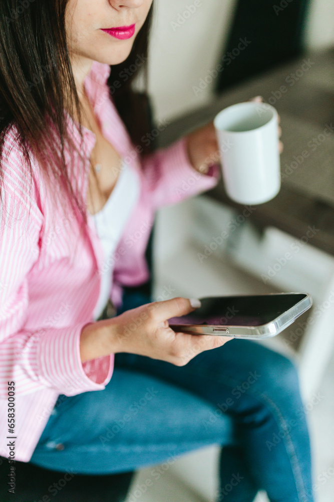 © Mila - A young girl with black long hair in a pink shirt and white top sits at a table with a phone and a white mug in a cozy room. A white mug and a phone in the hands of a girl. Morning business woman