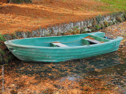 Abandoned turquoise fishing boat stands near the shore strewn with dry autumn orange leaves