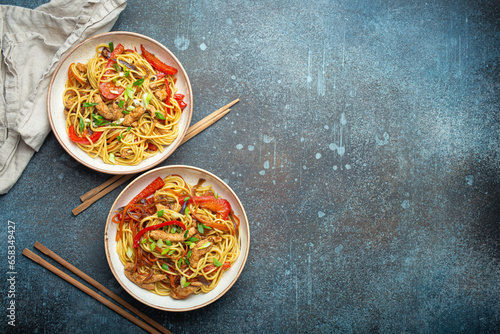 Two bowls with Chow Mein or Lo Mein, traditional Chinese stir fry noodles with meat and vegetables, served with chopsticks top view on rustic blue concrete background, space for text.