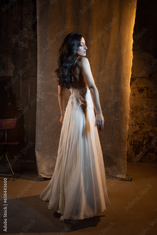 Elegant and beautiful brunette in a chic evening dress or wedding dress, white dress with a long flowing skirt to the floor. photo in dark colors on a textured wall, warm light