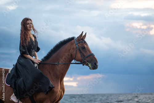 A beautiful young horsewoman in a black dress and with her hair down, riding a horse, portrait against the background of the evening sky, horseback riding in the open air
