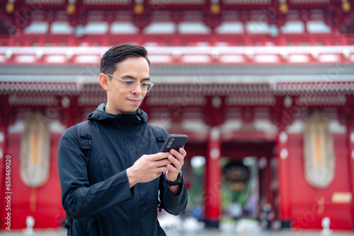 Hispanic tourist guy standing on blurred background of Japanese temple Sensoji  and sending text message via mobile phone during journey in Asakusa Tokyo, Japan photo