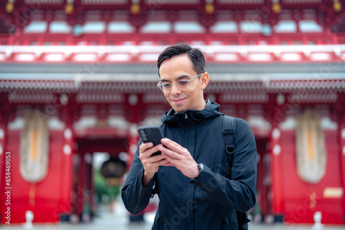 Hispanic tourist guy standing on blurred background of Japanese temple Sensoji  and sending text message via mobile phone during journey in Asakusa Tokyo, Japan photo