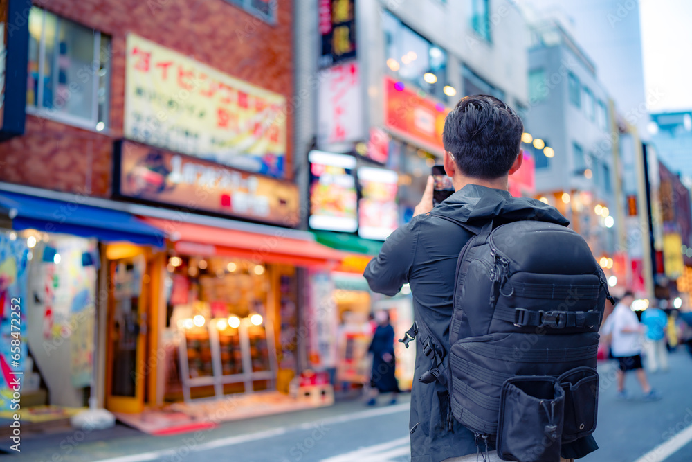 Back view of unrecognizable Hispanic male tourist with backpack taking picture on smartphone in bright Shinjuku neighborhood street of Tokyo city, Japan - obrazy, fototapety, plakaty 