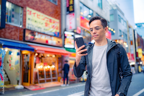 Focused young Hispanic male in casual clothes and eyeglasses walking on illuminated Shinjuku neighborhood in Tokyo while browsing smartphone in daylight, Japan photo