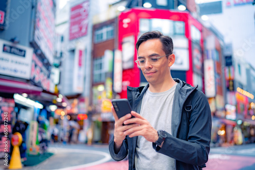 Happy young Hispanic male in casual clothes and eyeglasses walking on illuminated Shinjuku neighborhood in Tokyo while browsing smartphone in daylight, Japan photo