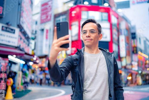 Focused young Hispanic male in casual clothes and eyeglasses taking selfie with smartphone on illuminated Shinjuku neighborhood in Tokyo in daylight, Japan photo