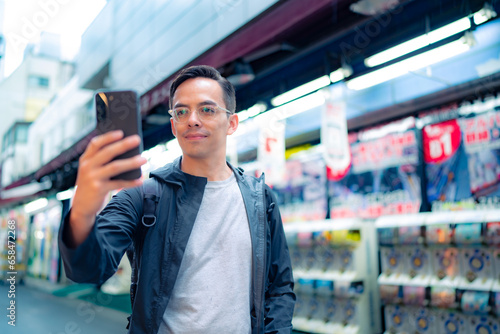 Focused young Hispanic male in casual clothes and eyeglasses taking selfie with smartphone on illuminated Shinjuku neighborhood in Tokyo in daylight, Japan photo