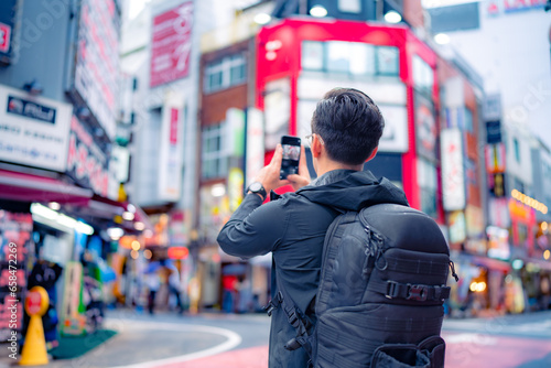 Back view of unrecognizable Hispanic male tourist with backpack taking picture on smartphone in bright Shinjuku neighborhood street of Tokyo city, Japan photo