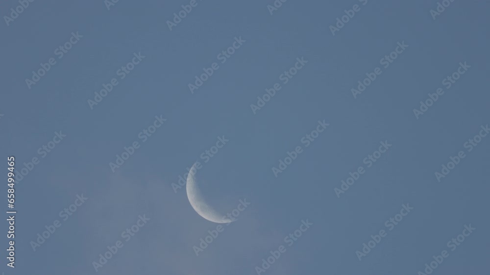 Crescent moon in blue sky during day with faint clouds wisping over Crescent moon in blue sky during day with faint clouds wisping over