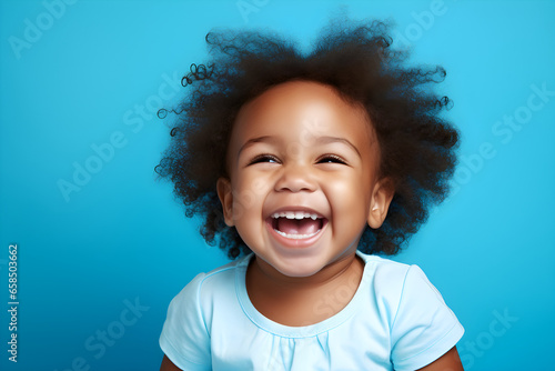Portrait of a cute African American baby girl wearing blue tee shirt laughing on bright blue background