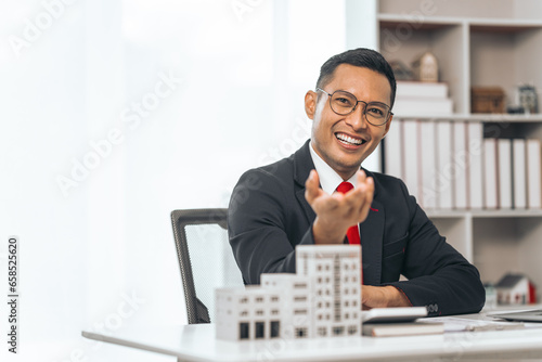 Agent presents contracts for property purchase or lease. Businessman asian indian people showcases miniature model home, signifying secure property insurance. Desk scene in a home sales office.