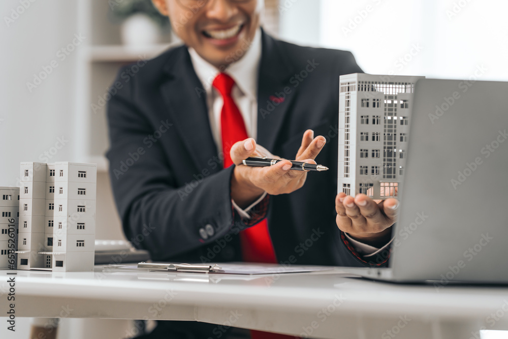 Agent presents contracts for property purchase or lease. Businessman asian indian people showcases miniature model home, signifying secure property insurance. Desk scene in a home sales office. Agent presents contracts for property purchase or lease. Businessman asian indian people showcases miniature model home, signifying secure property insurance. Desk scene in a home sales office.