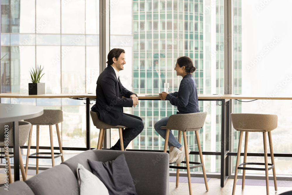 Positive Indian and Caucasian colleagues discussing cooperation, partnership terms. Happy confident diverse professional couple meeting in office coworking space, talking at large city window. Positive Indian and Caucasian colleagues discussing cooperation, partnership terms. Happy confident diverse professional couple meeting in office coworking space, talking at large city window.