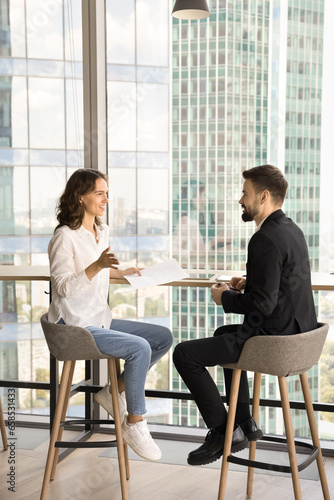Two young entrepreneurs discussing cooperation in co-working space, talking at window with urban downtown view background, smiling, enjoying networking, collaborating on project