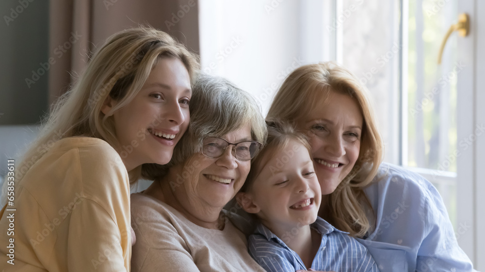 Happy cute female relatives posing for camera with toothy smiles, looking away, sitting close on home sofa, hugging with love, meeting due to 8 march, mothers day. Family portrait, banner shot