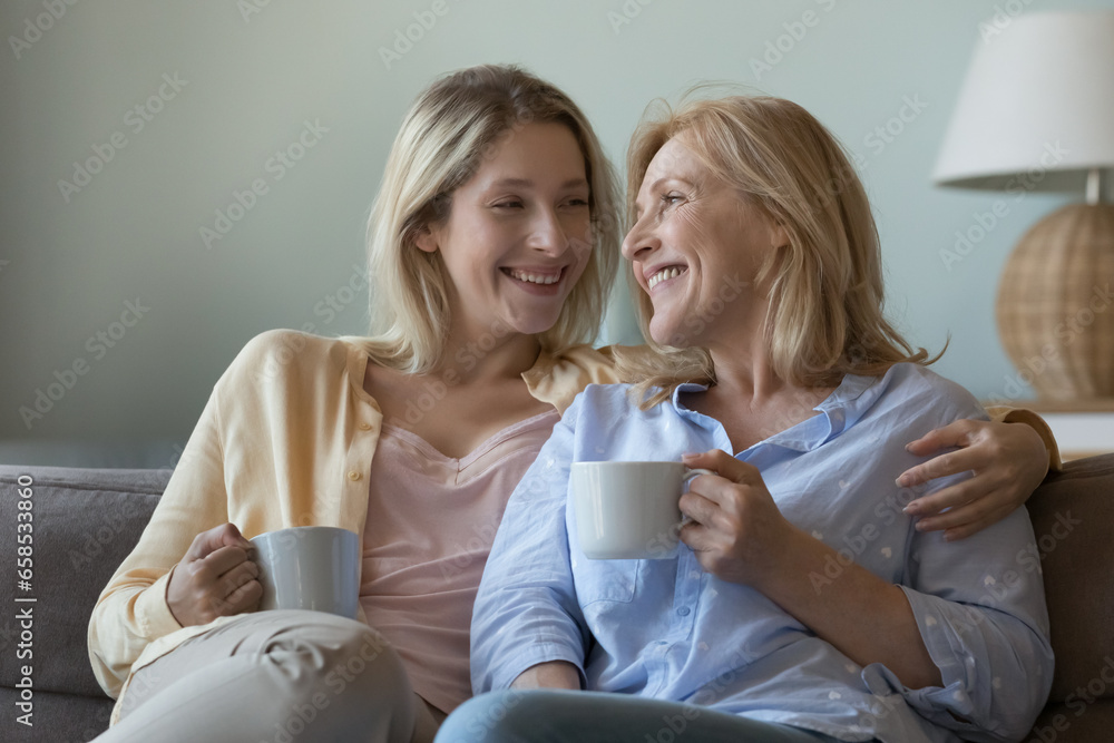 Cheerful loving young grownup daughter and happy mature mom drinking tea, coffee at home, holding cups, talking, laughing, hugging with love, tenderness, support, enjoying family leisure