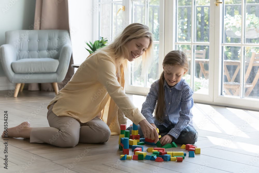 Happy young Caucasian mom and daughter kid playing on warm floor at home, constructing model from colorful toy blocks. Baby sitter, daycare teacher enjoying learning game with child - obrazy, fototapety, plakaty 