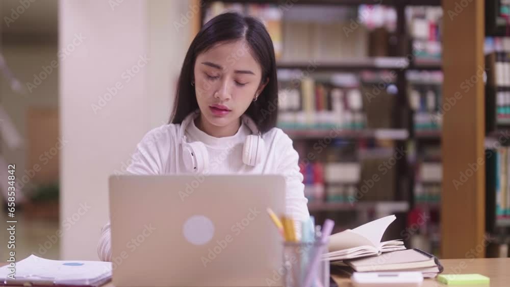 Concentrated Asian teen women student sitting at desk and using laptop for online learning class, and writing notes while preparing for exam in library campus. Concentrated Asian teen women student sitting at desk and using laptop for online learning class, and writing notes while preparing for exam in library campus.