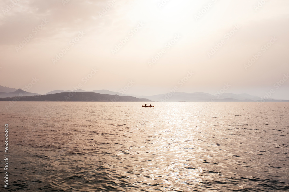 Misty mood seascape with fishermen boat in the middle of the sea and coastline in background under fog sunlight.