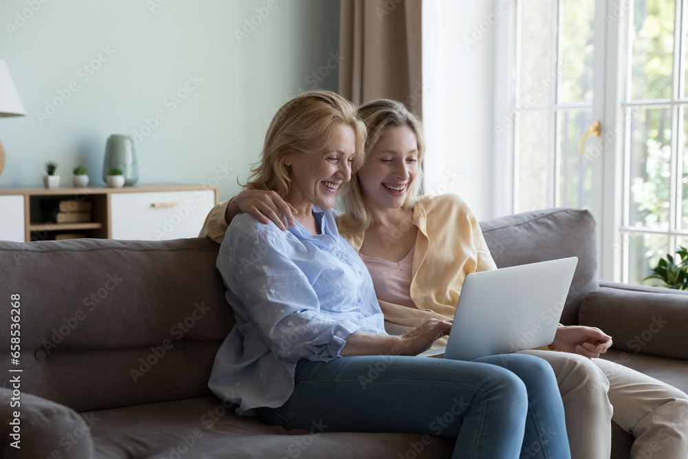 Happy pretty blonde senior mom and young adult daughter woman watching movie, series online broadcasting on laptop computer, laughing, talking on video call, enjoying wireless Internet communication Happy pretty blonde senior mom and young adult daughter woman watching movie, series online broadcasting on laptop computer, laughing, talking on video call, enjoying wireless Internet communication