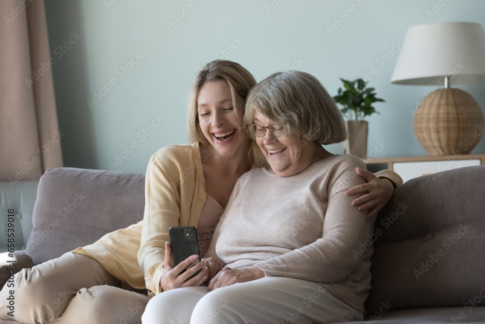 Happy pretty young adult granddaughter and elder grandma talking on video call on smartphone, taking selfie, holding mobile phone, sitting on couch together, hugging, laughing at gadget screen