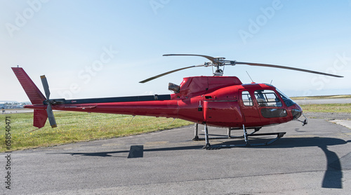 small red tourist helicopter sitting on the ground at an airport with a clear blue sky in the background and tarmac in the foreground