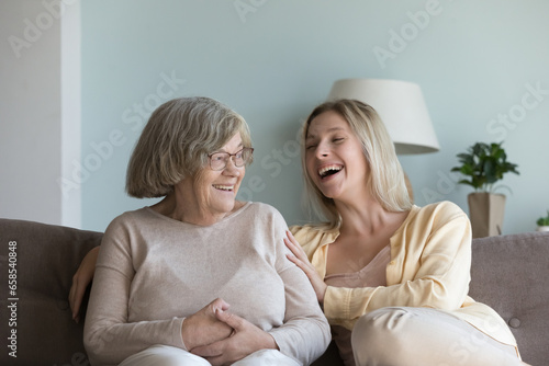 Cheerful pretty young adult granddaughter woman and elder grandma talking, chatting, laughing on home couch, enjoying family leisure time, warm close relationships, meeting © fizkes