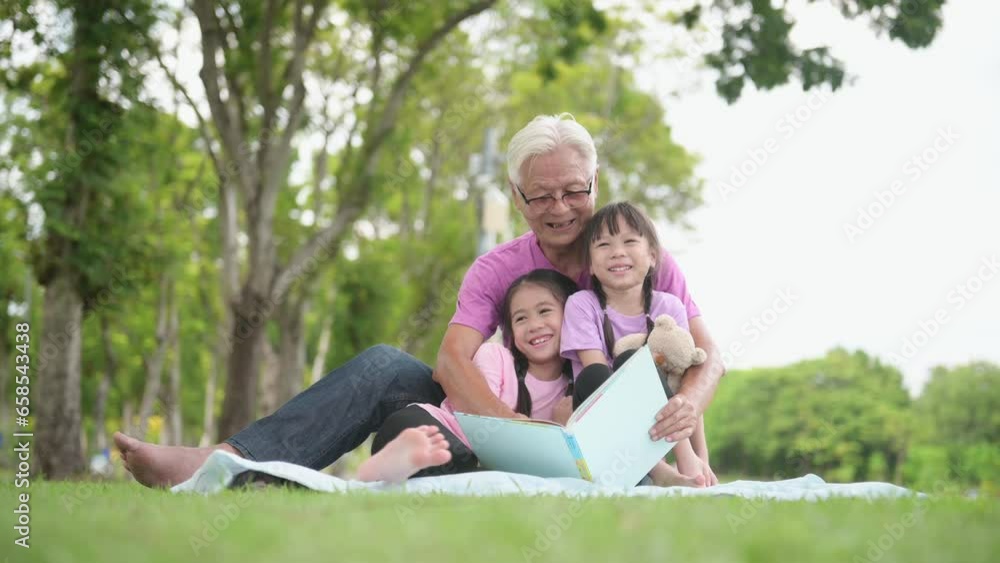 Happy Asian family children reading a book with her grandfather in the park Happy Asian family children reading a book with her grandfather in the park