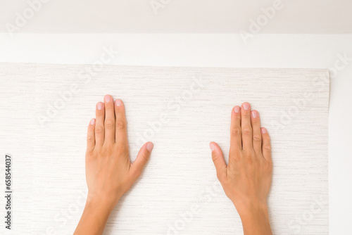 Young adult woman hands applying new light gray wallpaper on white wall. Closeup. Repair work of home. Point of view shot.
