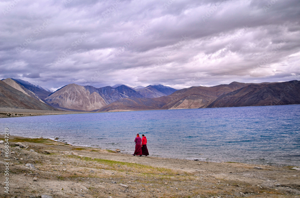 Two Buddhist monks taking a stroll on the banks of a lake with blue pristine water under a cloudy overcast morning with a backdrop of mighty Himalayan mountain range