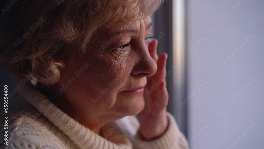An upset senior woman wiping tears, crying alone near the window, dealing with loneliness in her age An upset senior woman wiping tears, crying alone near the window, dealing with loneliness in her age