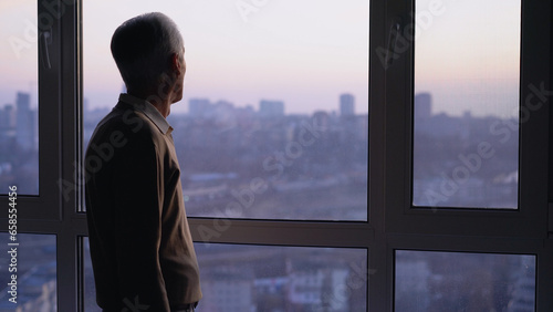 A lonely, thoughtful man in his 70s looking at a big window, waiting for relatives to arrive