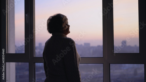 A lonely senior woman sadly looking out the window, missing her family at the retirement home © Synthex🇺🇦