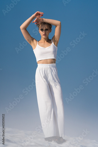 Authenticity slim blond woman with short hair dressed in white crop top and white pants. Caucasian ethnicity female looking at camera, posing through snow with light fog on sunny weather with blue sky