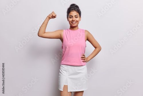 Photo of strong sporty woman raises arm and shows biceps motivates you for doing sport wears pink t shirt and skirt smiles gladfully isolated over white background. You can do it. Healthy lifestyle