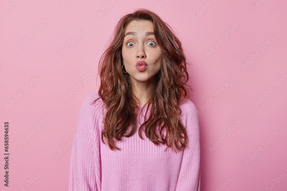 Fototapeta premium Horizontal shot of lovely dark haired young woman keeps lips folded stares surprised wants to kiss you dressed in knitted jumper isolated over pink background. Human facial expressions concept