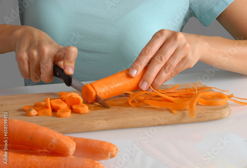 Woman slicing fresh carrots with a nkife on kitchen cooking table.
