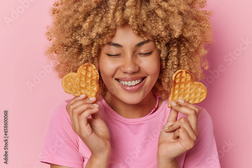 Fototapet Cheerful beautiful woman with curly hair holds two heart shaped waffles smiles broadly keeps eyes closed imagines how she eats this tasty food dressed casually isolated over pink background