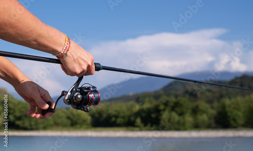 Spinning fishing. Female hands hold a spinning rod against the background of a mountain river, mountains and blue sky in summer on a sunny day, close-up.