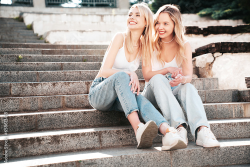 Two young beautiful smiling hipster female in trendy summer white t-shirt and jeans clothes. Carefree women posing in the street. Positive models having fun outdoors. Cheerful and happy. Sit at stairs